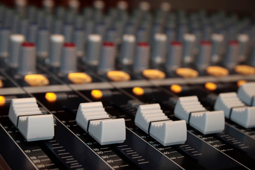Table de mixage audio avec rangées de faders gris et échelles numérotées de 0 à l'infini. Indicateurs lumineux jaunes en arrière-plan.
