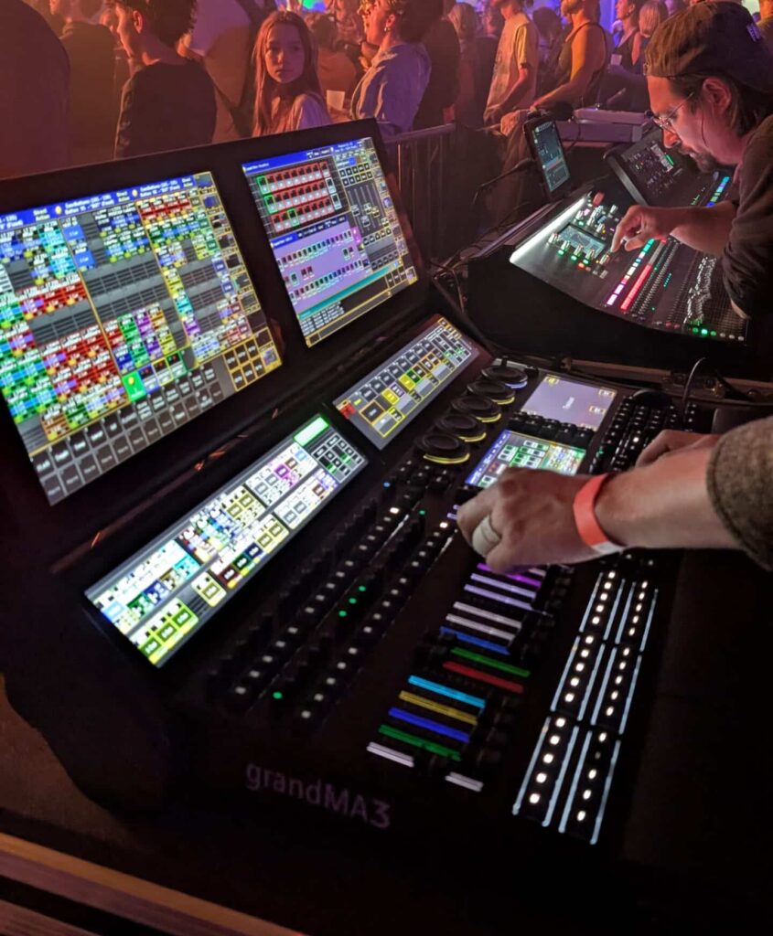 grandMA3 : Opérateur au pupitre d'éclairage de concert live. Opérateur grandMA3 gérant l'éclairage de concert. Mains sur la console, écrans multicolores, foule en arrière-plan sous des lumières vives.